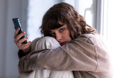 Pencerenin yanında otururken akıllı telefon kullanan kıvırcık bir kızın portresi.