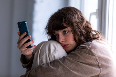 Pencerenin yanında otururken akıllı telefon kullanan kıvırcık bir kızın portresi.