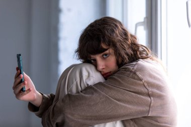 Kıvırcık kız pencerenin yanında otururken akıllı telefon kullanıyor..