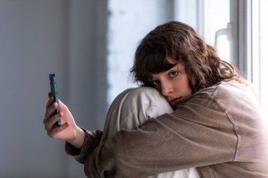 Kıvırcık kız pencerenin yanında otururken akıllı telefon kullanıyor..