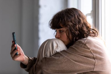 Kıvırcık kız pencerenin yanında otururken akıllı telefon kullanıyor..