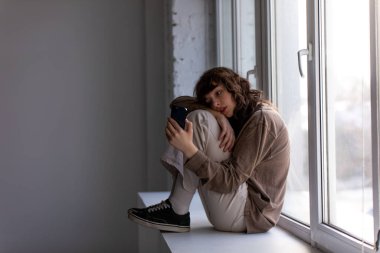 Elinde telefon olan kıvırcık, genç bir kadın pencere eşiğinde oturuyor..