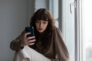 Elinde telefon olan kıvırcık, genç bir kadın pencere eşiğinde oturuyor..