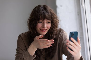 Kıvırcık genç kadın akıllı telefon kullanarak görüntülü konuşma yapıyor..