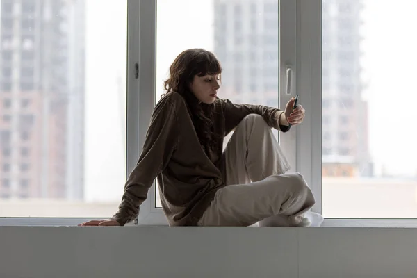 Young attractive woman uses smartphone while sitting on the windowsill by the window. — ストック写真