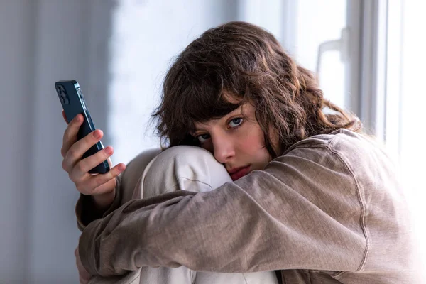 Portrait of a curly girl using a smartphone while sitting by the window. — ストック写真