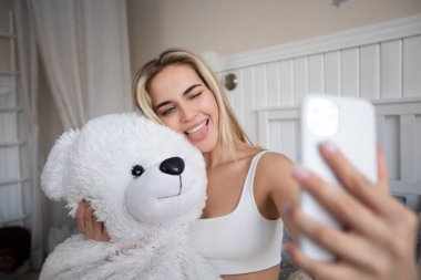 Güzel sarışın kadın yatakta oyuncak ayıyla selfie çekiyor..