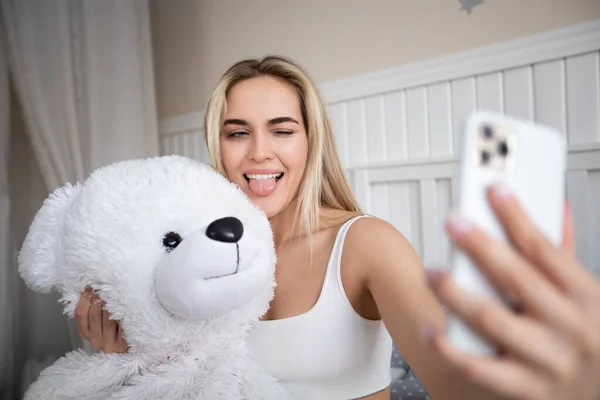 Beautiful blonde woman makes a selfie with a teddy bear sitting on the bed.