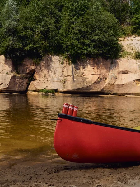 Cesis, Letônia - julho de 2021: vista para a canoa com três latas de Coca-Cola contra o rio Gauja e paredes verticais de areia no fundo. — Fotografia de Stock