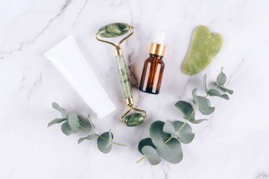 Yeşim bir yüz silindiri ve güzellik serumlu gua sha aleti beyaz mermer masa arkasında yüz kremi ve yüz kremi ile yatıyordu. Masaj terapisi için yüz masajı seti, spa. Model