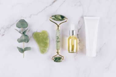 Yeşim yüz silindiri ve gua sha aleti ile düz yatıyordu güzellik serumu, yüz yağı ve yüz nemlendirici beyaz mermer masa arkasında. Masaj terapisi için yüz masajı seti, spa. Model