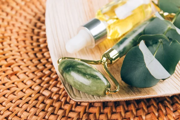 Doğal saman arka planında güzellik serumu yağı olan yeşim taşlı yüz silindiri yakın plan. Doğal taşlardan yapılan masaj terapisi için yüz masajı kiti, yaşlanma önleyici aletler.