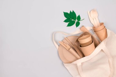 Eco-friendly tableware - kraft paper cups and containers in white cotton bag on gray background with copy space for text. Street food take away paper packaging - cups, plates, drinking straws