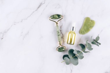 Yeşim bir yüz silindiri ve güzellik serumlu gua sha aleti ve beyaz mermer masa zemininde yüz yağıyla yatıyordu. Doğal taşlardan yapılan masaj terapisi için yüz masajı kiti.