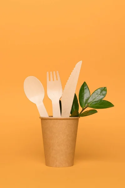 Natural wooden cutlery set made of natural bamboo in paper cup with green leaves - biodegradable or compostable cutlery concept. Vertical image on orange background. Environmental conservation concept