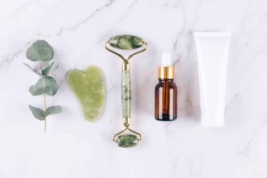 Yeşim yüz silindiri ve gua sha aracı, beyaz mermer masa arkasında yüz serumu ve kremi. Doğal taşlardan yapılan kaldırma terapisi için yüz masajı seti. Cilt bakımı yaşlanmayı önleyici aletler. Düz yatıyordu