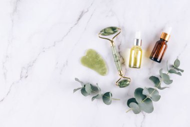 Yeşim yüz silindiri ve gua sha aracı, beyaz mermer masa zemininde yüz serumu veya organik yağ. Doğal taşlardan yapılan kaldırma terapisi için yüz masajı seti. Cilt bakımı yaşlanmayı önleyici aletler. Düz yatıyordu