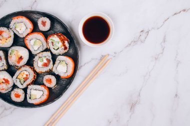 Suşi barı öğle yemeği menüsü, üst manzara. Somon, avokado ve beyaz mermer masa arkasında önlüklü yılan balıklı farklı suşi dürümleri. İnternetten sipariş edilen yemekler, paket servis konsepti. Seçici odak