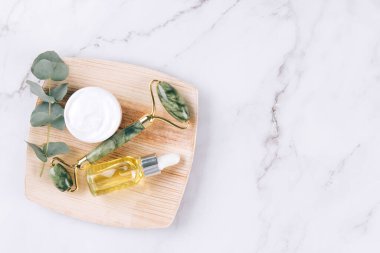Yeşim yüz silindiri ve gua sha aleti ile güzellik serumu, yüz yağı ve yüz nemlendirici beyaz mermer masa arkasında. Masaj terapisi için yüz masajı seti, spa, modelleme. Üst görünüm