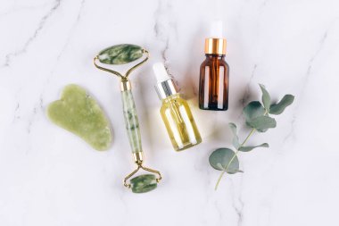 Yeşim yüz silindiri ve gua sha aracı, beyaz mermer masa zemininde yüz serumu veya organik yağ. Doğal taşlardan yapılan kaldırma terapisi için yüz masajı seti. Cilt bakımı yaşlanmayı önleyici aletler. Düz yatıyordu