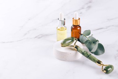 Yeşim yüz silindiri ve gua sha aleti ile güzellik serumu, yüz yağı ve yüz nemlendirici beyaz mermer masa arkasında. Masaj terapisi için yüz masajı seti, spa, modelleme.