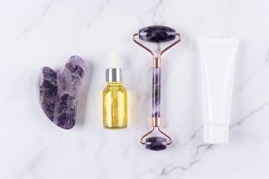 Yüz masajı kiti - ametist gua sha aracı ve yüz silindirli güzellik yüz serumu, kozmetik yağ ve beyaz mermer arka planda yüz nemlendirici kremi. Yüz silindiri, doğal taşlardan yapılmış masaj aleti. Seçici odak
