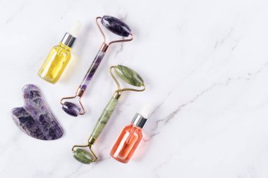Yüz masajı kiti - ametist ve yeşim taşıma aleti ve yüz silindirli güzellik yüz serumu, kozmetik yağ ve beyaz mermer arka planda yüz nemlendirici kremi. Yüz silindiri, doğal taşlardan yapılmış masaj aleti.