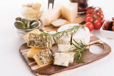Tahta peynir tahtası, zeytin, kiraz domatesleri ve beyaz masa arkasında krakerlerle servis edilen peynir seçimi. Aperatif için peynir tabağı - Brie, mavi peynir, gorgonzola, camembert 