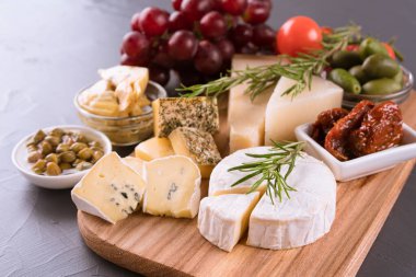 Peynirli şarküteri tahtası. Antipasti çeşitleri - brie, mavi peynir, gorgonzola, camembert, zeytin, kiraz ve güneşte kurutulmuş domates, yağlı enginar, kapari, üzüm. Gri taş arka planda peynir tabağı