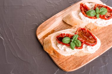 Ya ricotta peynirli tost ya da ahşap kesim tahtasında fesleğenle kaplanmış güneşte kurutulmuş domates. Gri masa arkası. İtalyan mezesi.