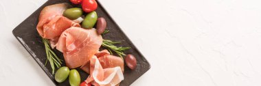 Banner, siyah şarküteri tahtasında zeytin ve bebek domateslerle servis edilen beyaz beton masa zemininde fotokopi alanı ile üst manzara. Geleneksel İtalyan antipastasıyla siyah tabak, İtalyan aperatifi. Seçici odak