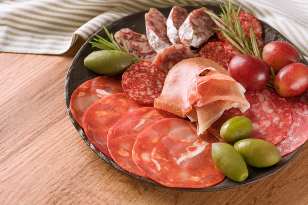 Farklı et aperatifleri, farklı türde sosisli şarküteri tabağı - salam, bresaola, zeytin, güllük ve taze üzümle servis edilen proscuitto. Tahta masada geleneksel İtalyan antipasti ile siyah tabak.