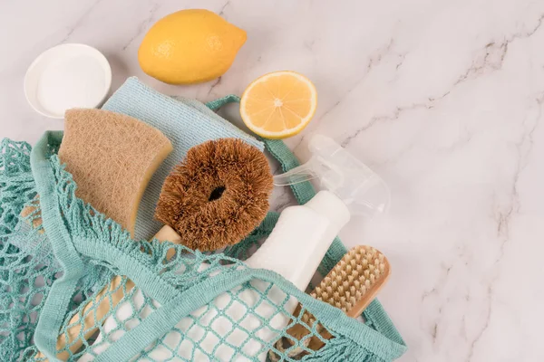Green household products in net bag on white marble background with copy space. Bamboo brush, organic dishcloth, lemon, baking soda, citric acid and spray bottle with cleaning liquid