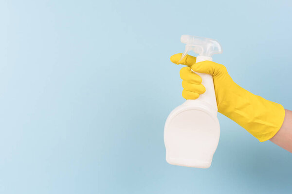 Hand in yellow protective rubber glove holding spray bottle with detergent against blue background with copy space. Cleaning product for different kitchen or bathroom surfaces. Bleach. Mockup