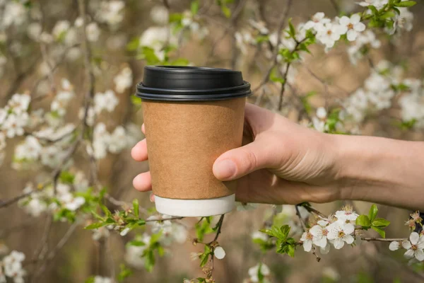 Arka plan olarak çiçek açan kiraz ağacına karşı Kraft kağıt kahve fincanı tutan genç bir kadın. Bahar kahvesi konsepti. Kraft kağıt kahve fincanı, plastik kapaklı.