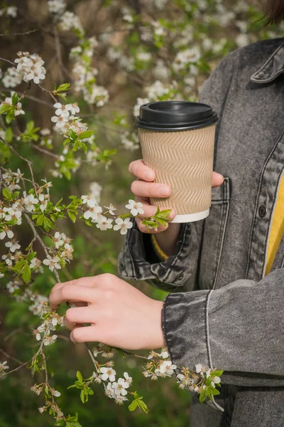 Arka plan olarak çiçek açan kiraz ağacına karşı Kraft kağıt kahve fincanı tutan genç bir kadın. Bahar kahvesi konsepti. Dikey resim