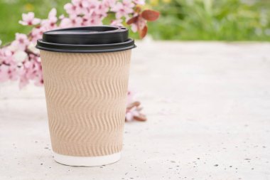 Arka plan olarak bulanık sakura ile beton masa üzerinde plastik kapaklı kahverengi kraft kağıt kahve fincanı görüntüsü. Kahveni götür. Şablon, boşluğu kopyala