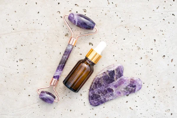 Yüz masajı kiti ile düz kompozisyon - ametist gua sha aracı ve taş zeminde güzellik yüz serumu veya kozmetik yağ ile yüz silindiri. Model görüntü, cilt bakımı konsepti 