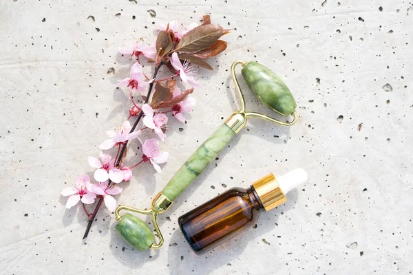 Taş beton zemin üzerine yüz masajı seti. Yüzünde güzellik serumu ya da kozmetik ve çiçek açan sakura dalı olan bir jade gua sha silindiri. Doğal taş yüz masajcısı, model. 