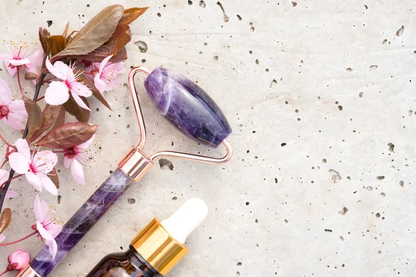 Yüz masajı kitinin yakın görüntüsü - ametist gua sha yüz silindiri ile güzellik yüz serumu ya da taş beton arka planda çiçek açan sakura dallı kozmetik yağ. Doğal taş yüz masaj aleti, boşluğu kopyala