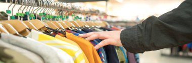Askıda erkek eli kıyafetleri olan pankart, alış veriş yapan adam. Moda mağazasında yeni moda koleksiyonu. Kıyafet kiralama ya da satış konsepti. Kıyafetler bulanık depo arka planına karşı fotokopi alanı ile oyalanır. Seçici odak