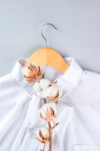 White organic cotton shirt on wooden hangers with cotton flowers over gray background with copy space. Conscious and environmentally friendly consupmtion - new modern trends in shopping. Slow fashion concept