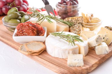 Beyaz masa arkasında çeşitli peynirler, zeytin, kurutulmuş domates ve otlarla servis edilen şarküteri peynir tahtası. Aperatif için peynir tabağı - Brie, dorblu, gorgonzola, camembert ve parmesan peyniri