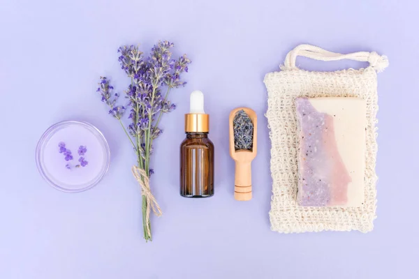 Natural herbal self-care products. Natural lavender cosmetics for home beauty treatments  - cosmetic lavender oil or face serum, organic soap and body lotion over purple background. Flat lay style