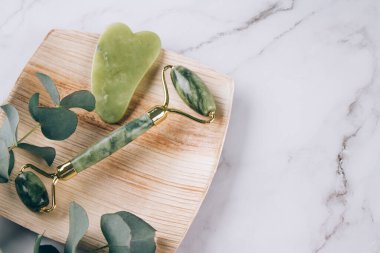 Yüz masajı destesi, yeşil yüz silindiri ve beyaz mermer masa zemininde gua sha kazıyıcı. Doğal taşlardan yapılan kaldırma terapisi için yüz masajı seti. Cilt bakım aletleri. Seçici odak