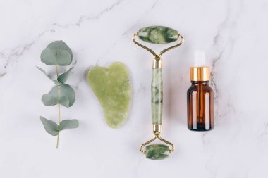 Jade facial roller and gua sha scraper, face serum or organic cosmetic oil over white marble table. Facial massage bundle for lifting therapy . Skin care anti-aging tools. Flat lay style