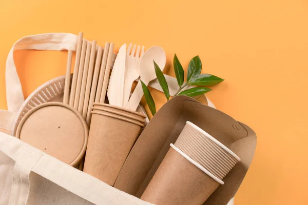 Kraft paper food cups, containers, wooden cutlery set, drinking straws in white cotton bag over orange background. Street food take away paper utensils. Selective focus