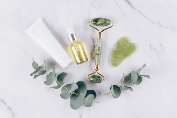 Jade facial roller and gua sha scraper, face cosmetic oil and face cream or cleanser over white marble table. Facial massage bundle for lifting therapy made from natural stones. Flat lay style