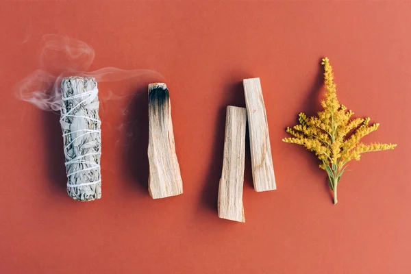 Burning incense. White sage, palo santo sticks  and fragrant herbs in a row over orange brown background. Bundle for meditation and room fumigation. Selective focus. Flat lay style