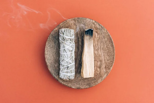 Burning incense bundle - white sage and palo santo stick with smoke on wooden stand over orange brown background. Holy wood sticks for meditation and room fumigation. Selective focus